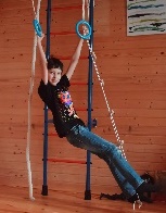 Boy hanging from blue rings and feet perched on rope swing. A ladder is the background of his home play space. 